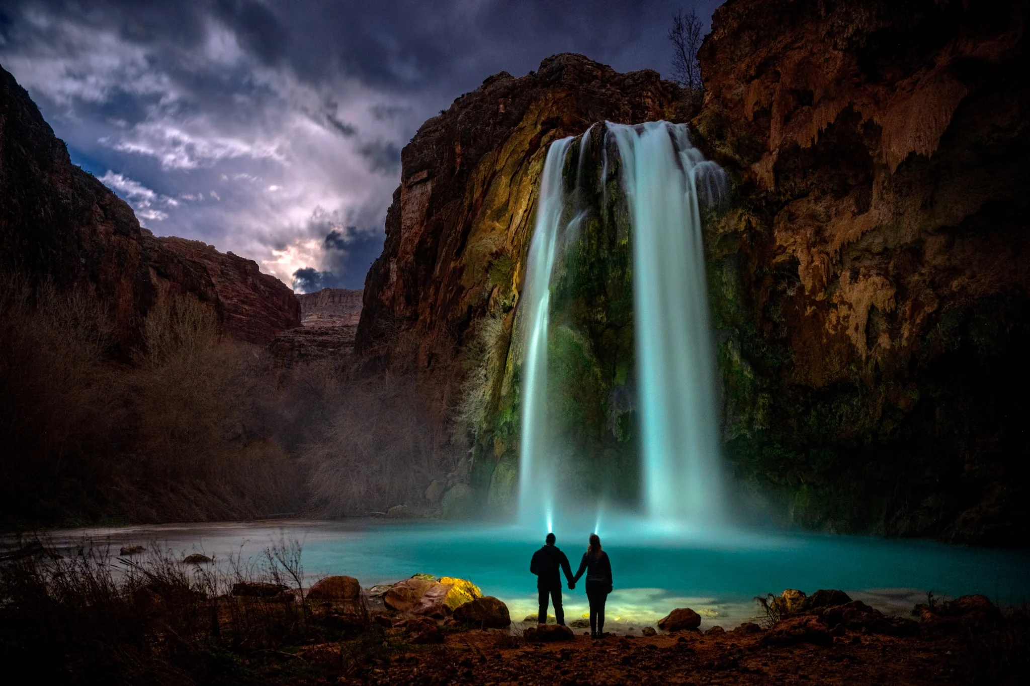 Two people holding hands before an illuminated waterfall beneath a dramatic night sky
